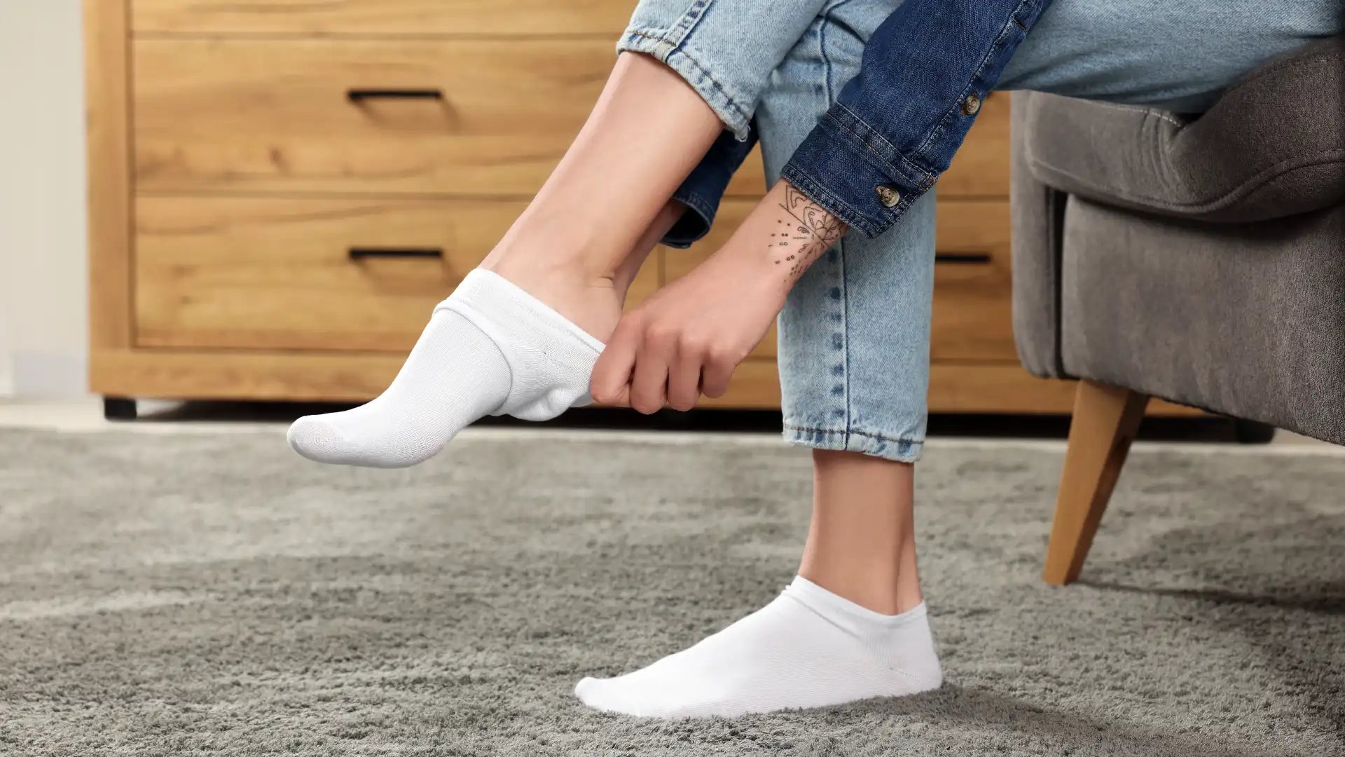 Une femme en jean assise dans un fauteuil gris enfile des mini-socquettes en bambou blanches pour aller travailler.